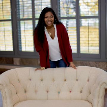 woman standing in white t-shirt and maroon cardigan behind a couch smiling