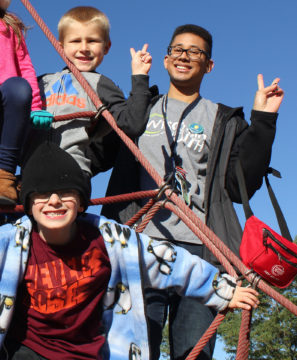 Two children and man standing on playground equipment smiling and holding peace signs with their fingers