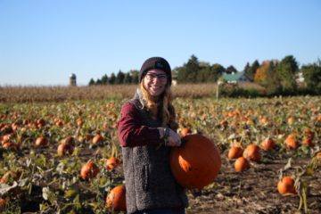woman wearing knit hat and vest standing in a pumpkin patch holding a pumpkin