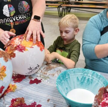 boy wearing green shirt with blonde hair sitting at table working on a paper mache project with two staff women on either side of him